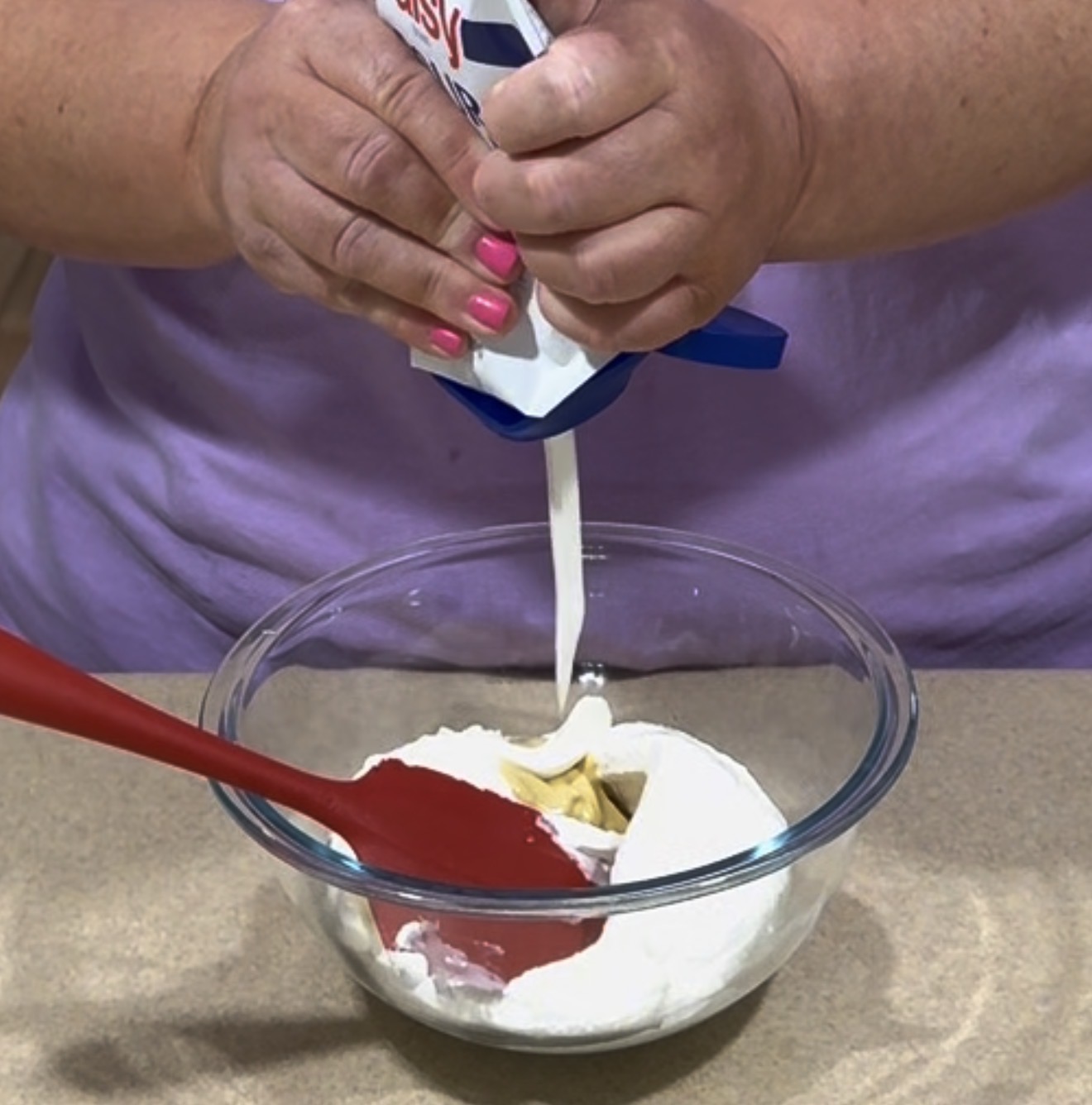 A person with pink nail polish pours cream from a carton into a glass bowl containing white powder and a stick of butter, using a red spatula, on a kitchen counter.