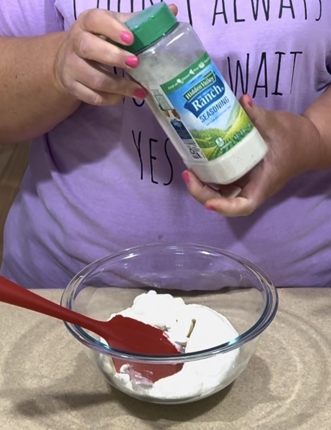 A person holding a container of ranch seasoning over a glass bowl filled with a white mixture, stirring with a red spatula. The person has pink nail polish and is wearing a lavender shirt with black text.