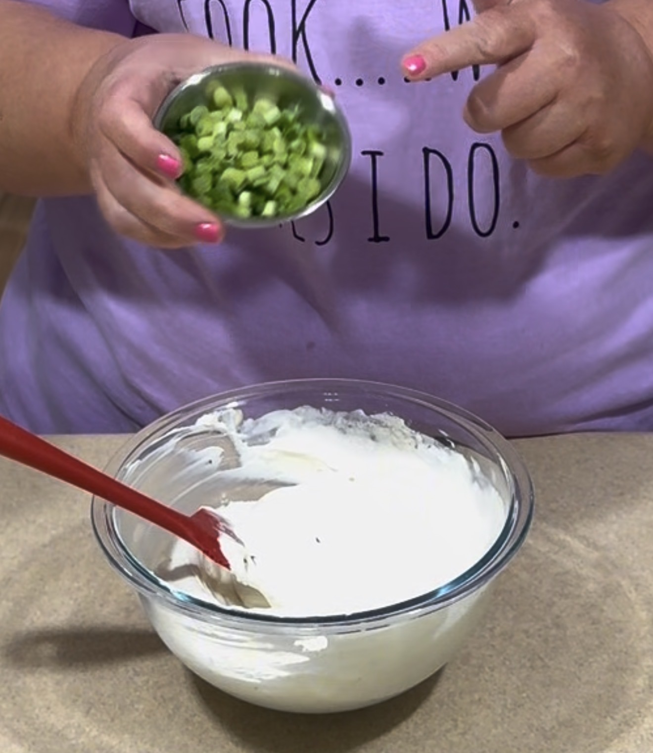 A person holding a small bowl of chopped green onions over a mixing bowl of creamy white mixture. A red spatula is in the bowl. The person is wearing a light purple shirt with black text.