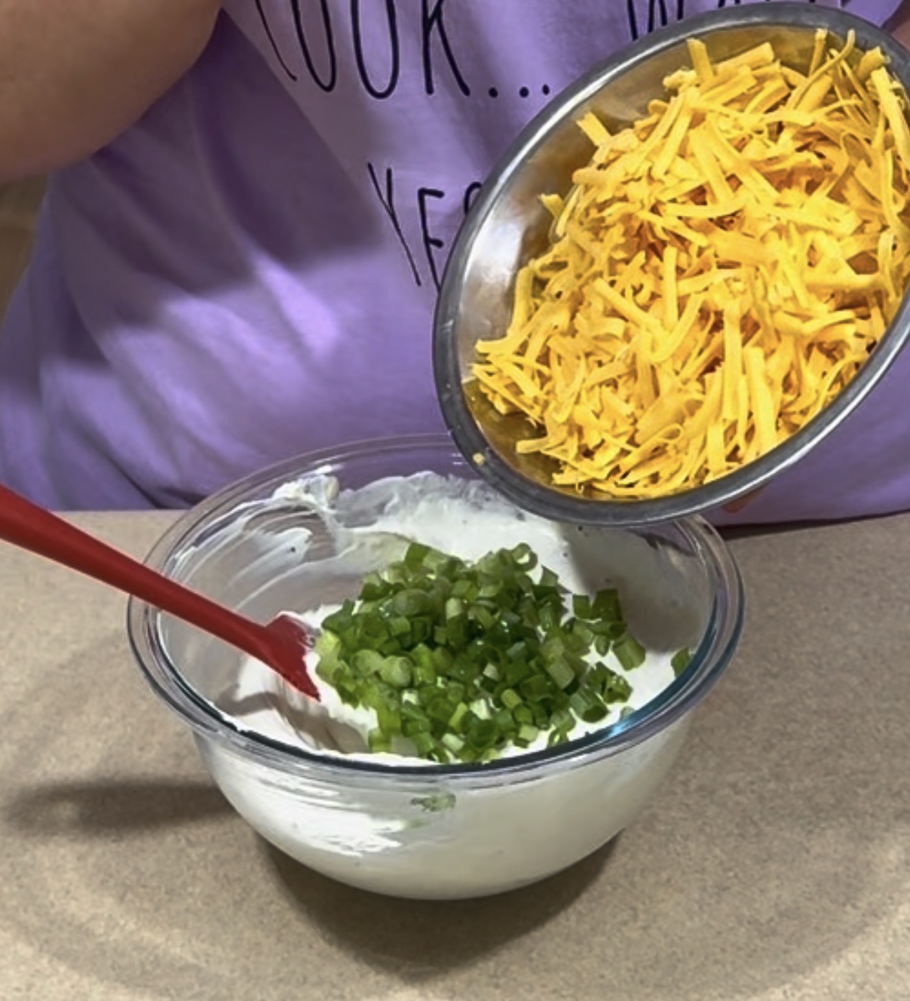 A person adds shredded cheddar cheese from a metal bowl into a glass bowl with chopped green onions and a creamy white mixture, stirring with a red spatula.