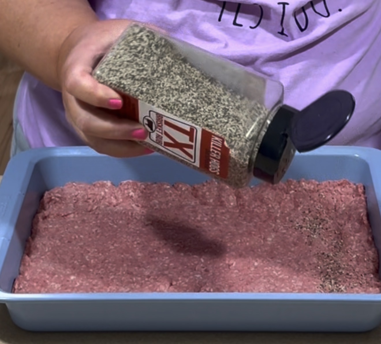 A person sprinkles black pepper from a large container onto a layer of raw ground meat in a blue baking dish. The person is wearing a light purple shirt with partial text visible.