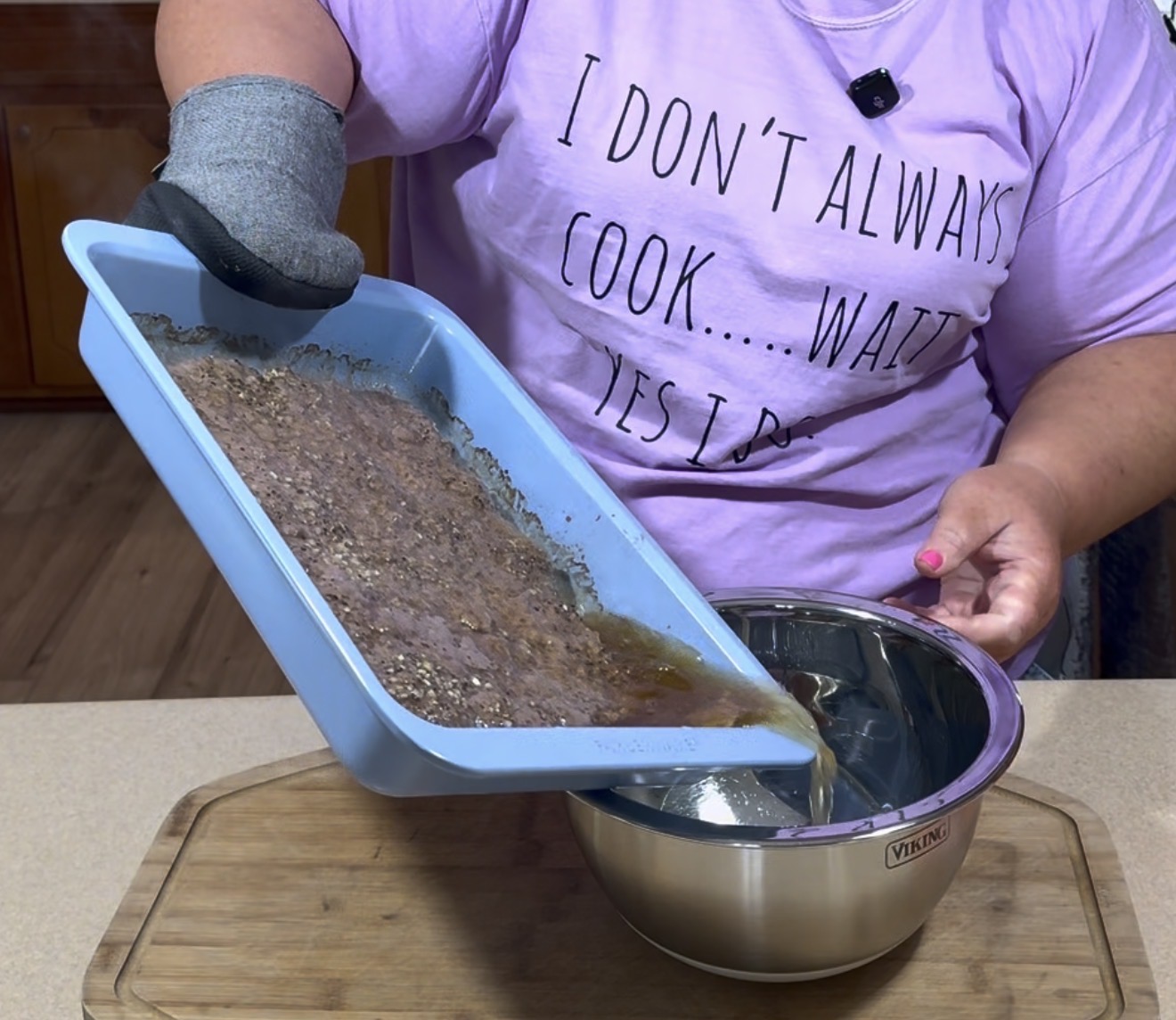 A person wearing an oven mitt and a shirt that says “I don’t always cook…wait, yes I do” pours liquid from a blue baking dish filled with seeds into a metal bowl on a kitchen counter.