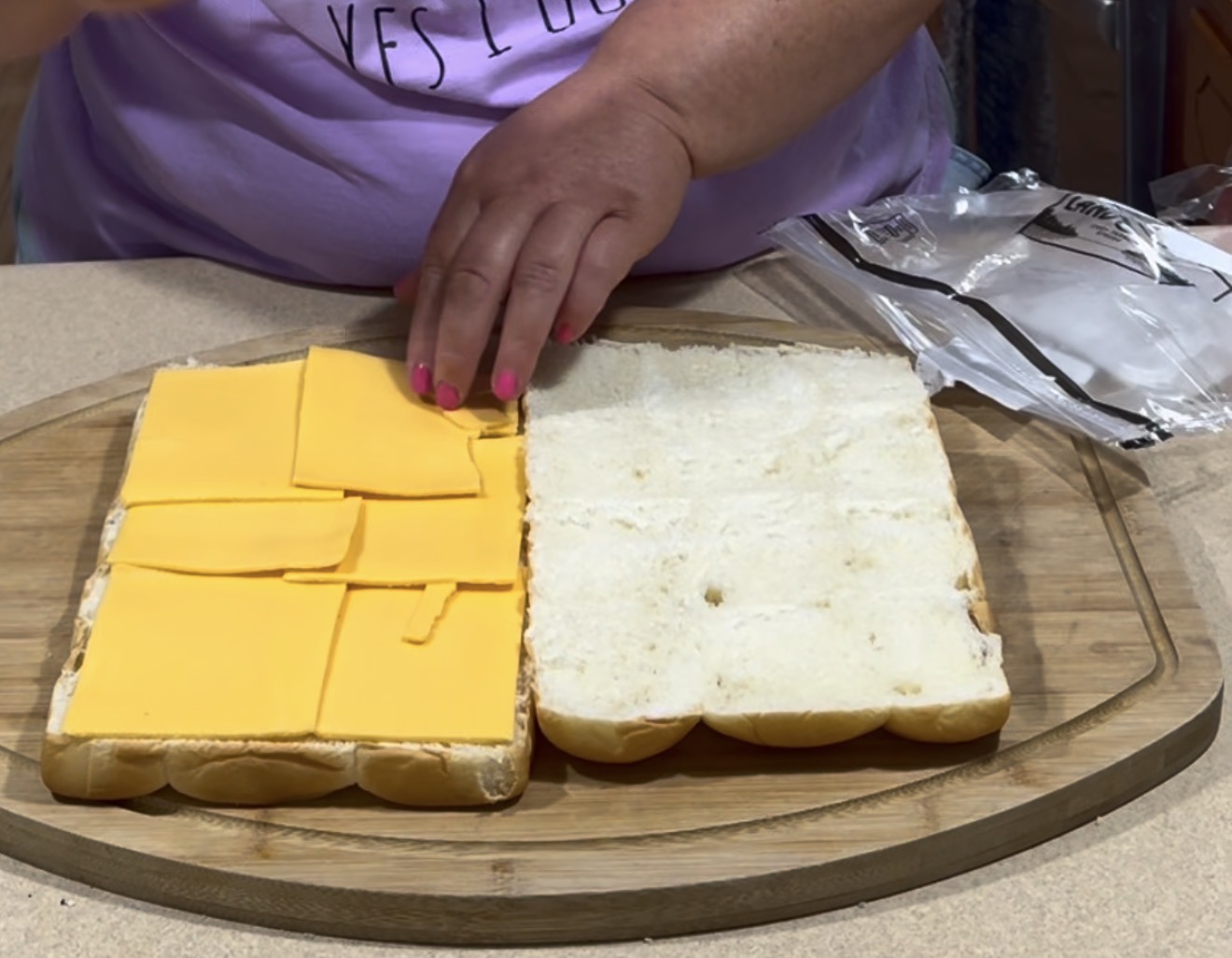 A person assembles a sandwich, placing slices of yellow cheese on one half of a large loaf of white sandwich bread, with an open plastic wrapper nearby on a wooden cutting board.