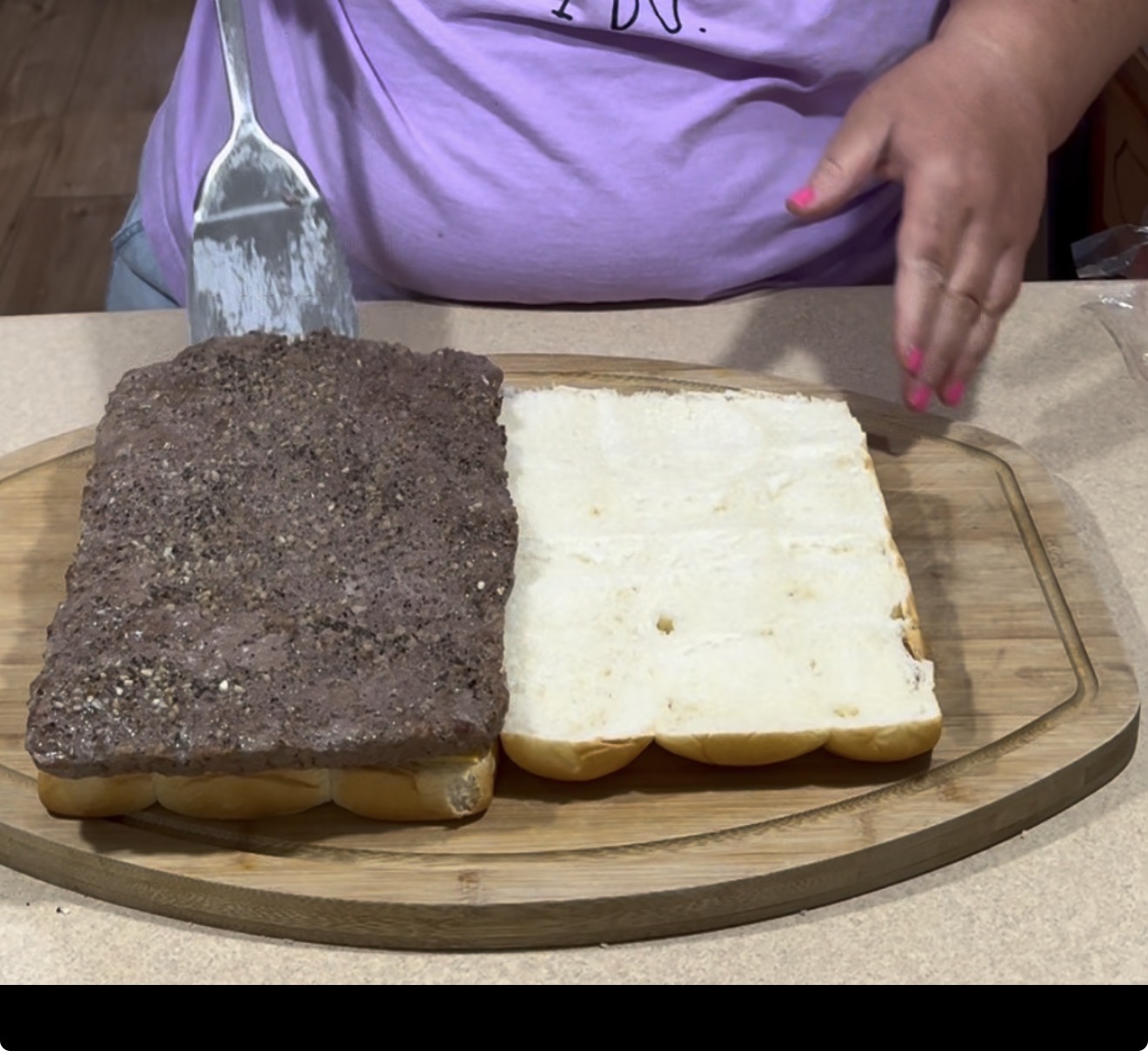 A person wearing a purple shirt spreads a dark, seasoned meat mixture onto a split loaf of white bread on a wooden cutting board.