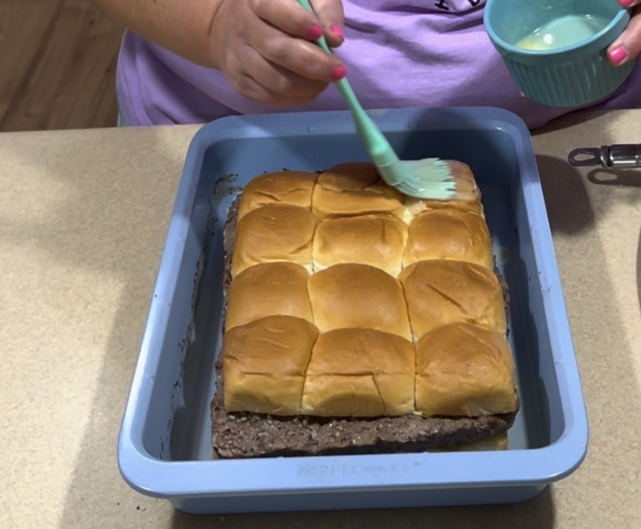 A person brushes melted butter onto a batch of baked slider buns that sit on top of a large cooked hamburger patty in a blue baking dish. The person holds a small bowl and wears a lavender shirt.