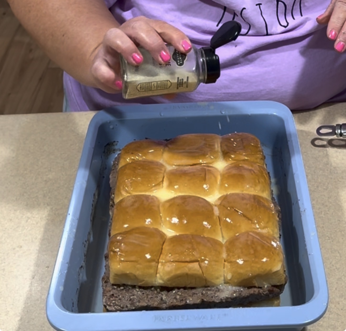 A person with pink nail polish sprinkles seasoning from a jar onto a tray of baked slider sandwiches in a blue baking dish.