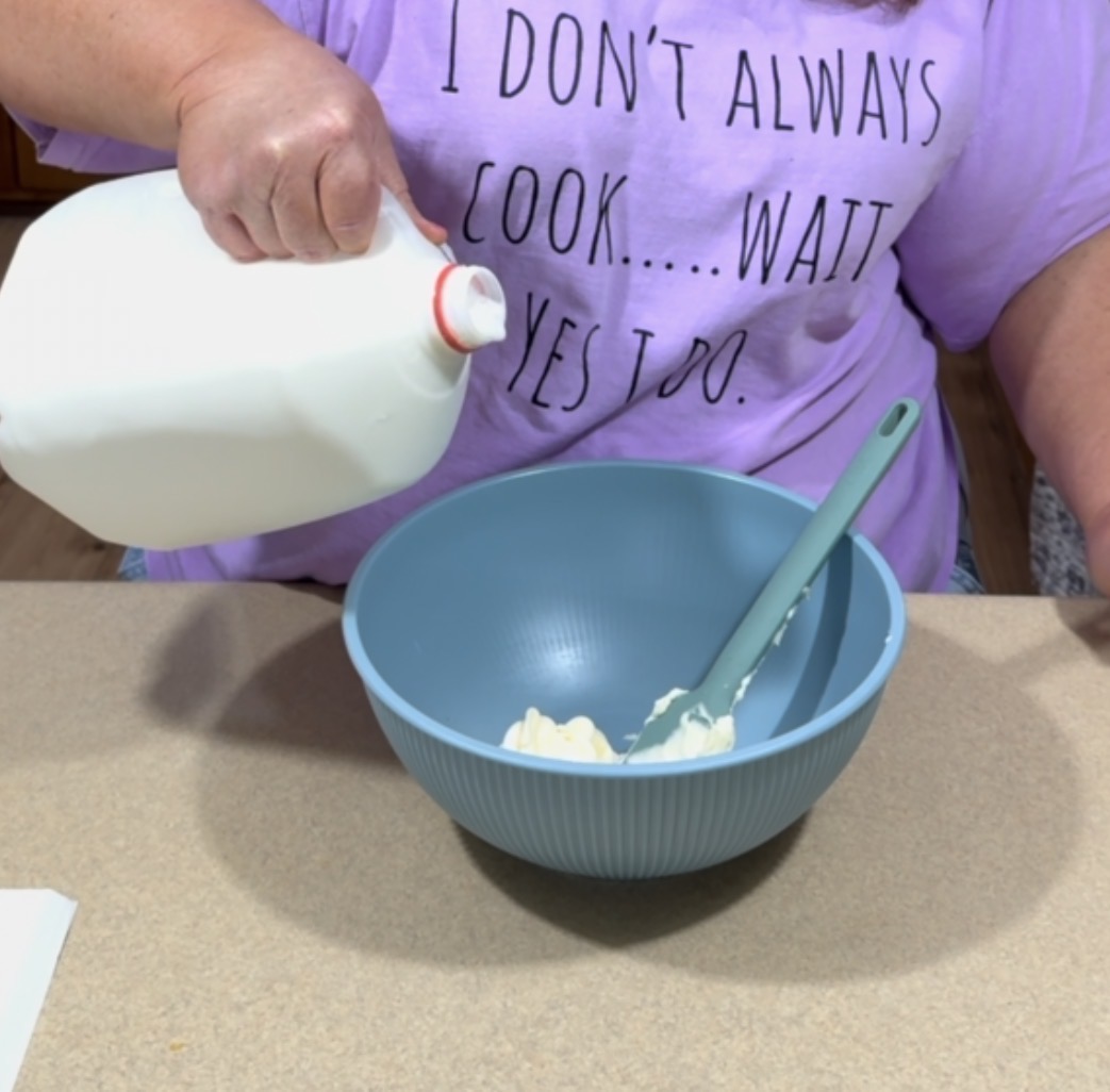 A person in a purple shirt that says “I DON’T ALWAYS COOK… WAIT, YES I DO.” pours milk from a jug into a blue mixing bowl on a counter; a green spatula rests in the bowl.