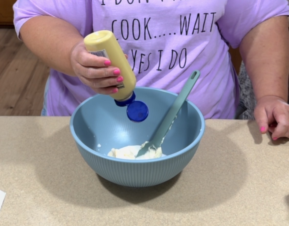 A person in a purple shirt squeezes mustard into a blue mixing bowl containing a white substance, possibly mayonnaise or cream cheese, while holding a spatula.