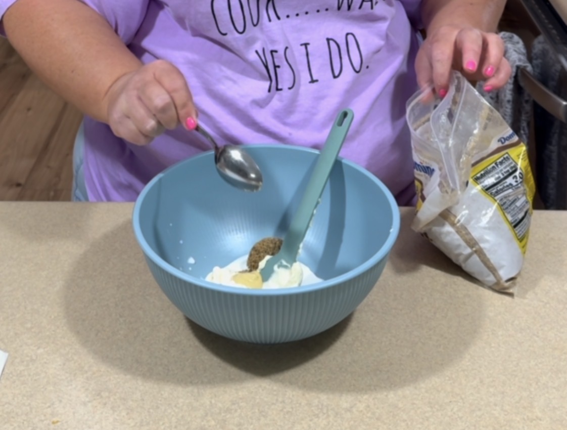 A person wearing a lavender shirt holds a spoon over a blue mixing bowl filled with ingredients while adding brown sugar from a bag. A green spatula is in the bowl on a kitchen counter.