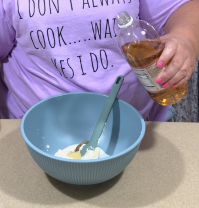 A person wearing a lavender shirt that says I dont always cook... wait, yes I do. is pouring liquid from a bottle into a blue mixing bowl with a spoon and white ingredients inside.