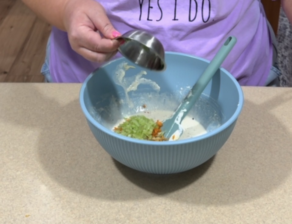 A person pours chopped green vegetables from a metal measuring cup into a blue mixing bowl with other ingredients. A teal spatula rests in the bowl. The person is wearing a purple shirt that reads YES I DO.