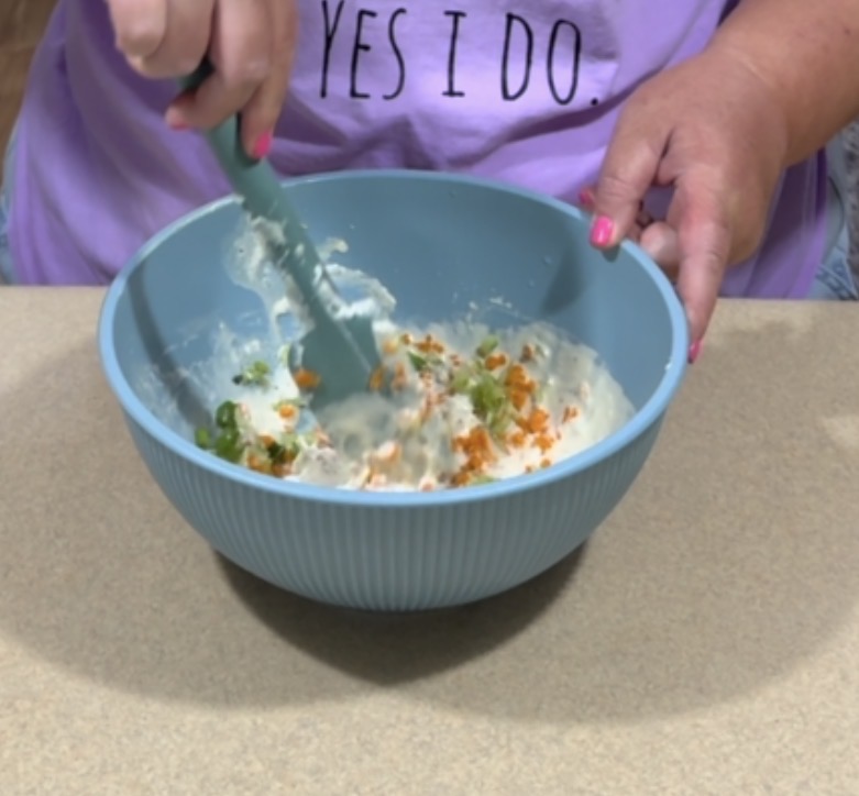 A person stirs a mixture with chopped green onions and orange spices in a blue bowl. They have pink nail polish and wear a purple shirt that says YES I DO.