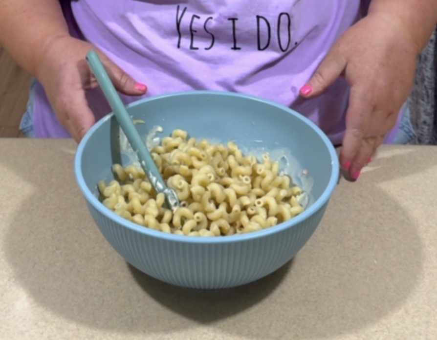A person wearing a lavender shirt with the words YES I DO. holds a blue bowl filled with cooked pasta and a mixing spoon, standing at a light-colored countertop.