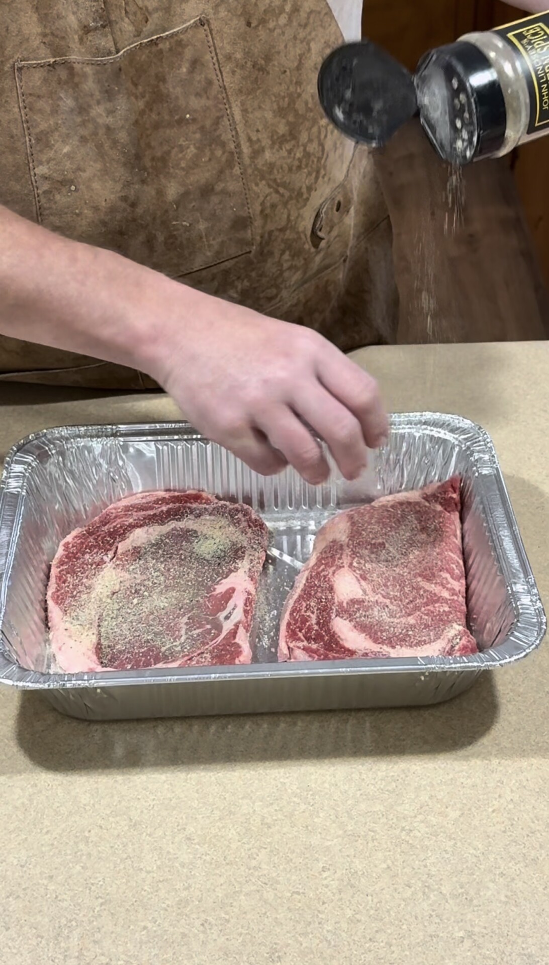 A person seasons two raw ribeye steaks with salt and pepper in a disposable aluminum tray on a countertop.