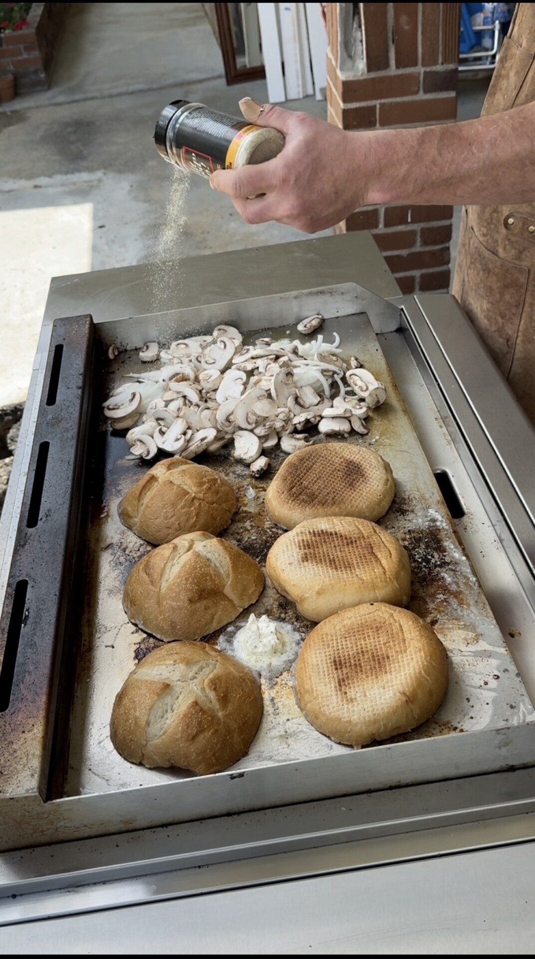 A person sprinkles seasoning over sliced mushrooms cooking on a flat top grill, alongside four round sandwich rolls and a pat of melting butter.