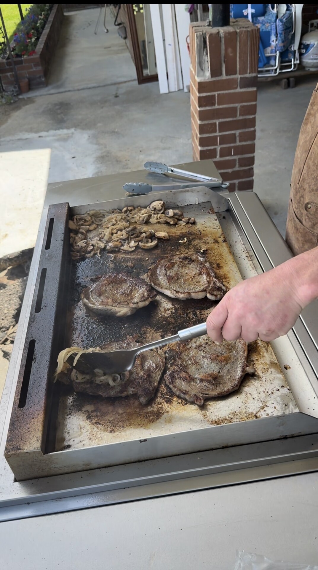 A person flips steaks and mushrooms on a large outdoor griddle with tongs. The food is sizzling and browned, and there is a brick pillar and patio area in the background.