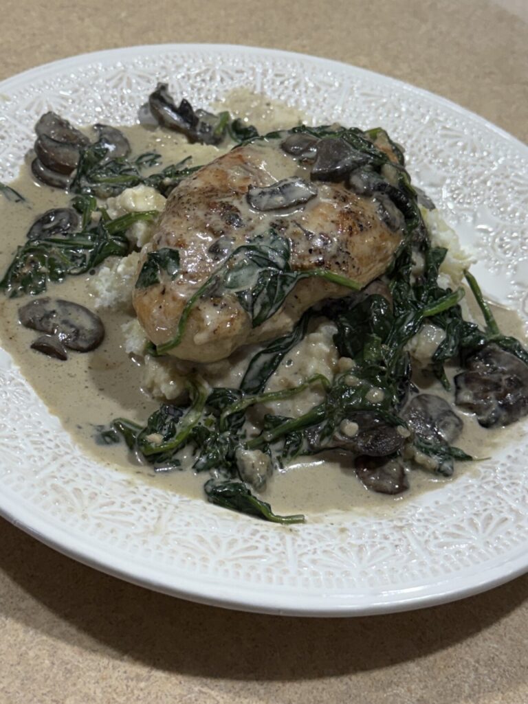 A plate of creamy chicken topped with sautéed spinach and mushrooms, served over mashed potatoes, on a decorative white plate sitting on a tan countertop.
