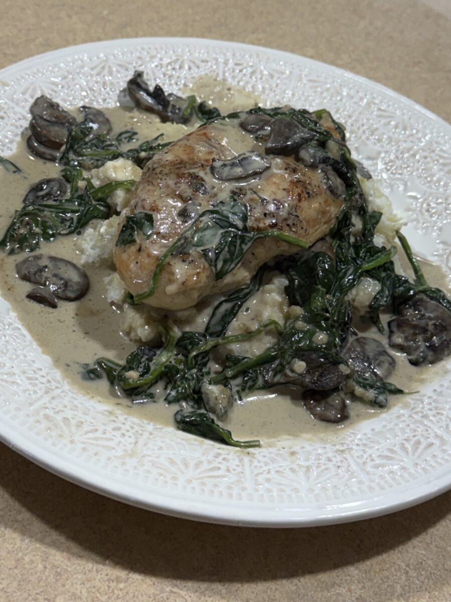 A plate of creamy chicken topped with sautéed spinach and mushrooms, served over mashed potatoes, on a decorative white plate sitting on a tan countertop.