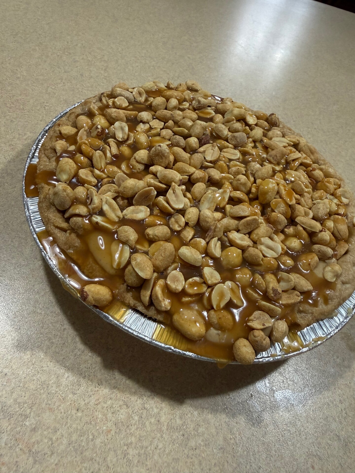 A pie in a foil pan topped with caramel sauce and a generous layer of whole peanuts sits on a beige countertop.