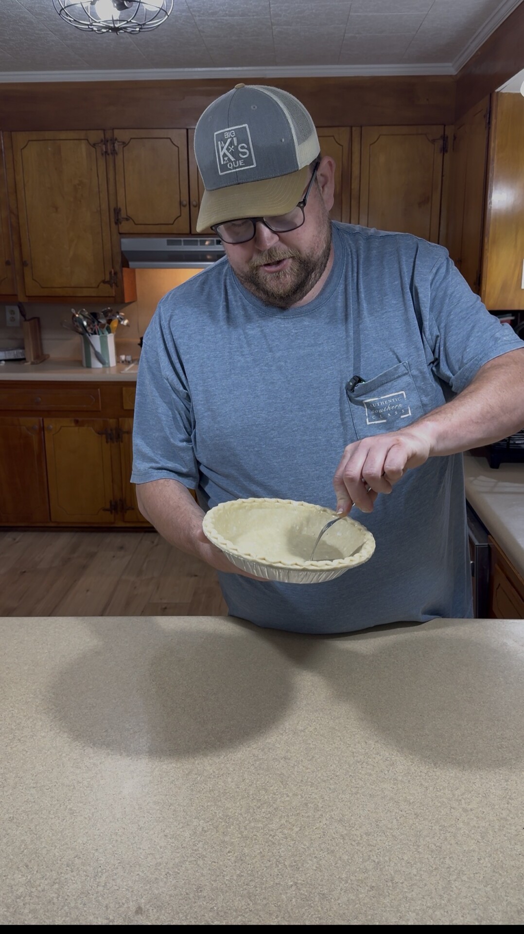 A man wearing a gray cap and blue shirt stands in a kitchen, poking holes in a raw pie crust with a fork over a countertop. Wooden cabinets and kitchen utensils are visible in the background.