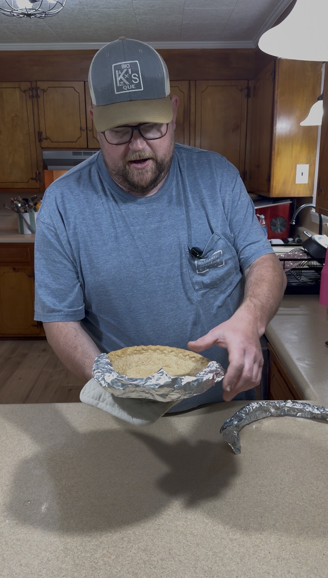 A man wearing glasses, a cap, and a blue shirt holds a pie with a foil crust shield in a kitchen. The kitchen has wooden cabinets and utensils in the background.