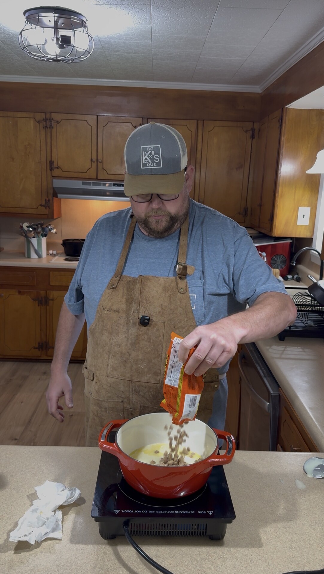 A man wearing a cap, glasses, and an apron pours a packet of chocolate chips into a red pot on a kitchen counter, with wooden cabinets and various kitchen items in the background.