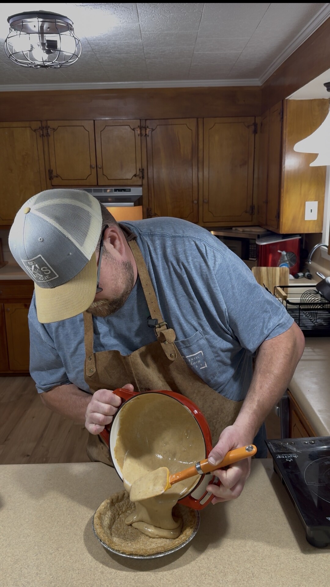 A man wearing a cap, glasses, and an apron pours cake batter from a mixing bowl into a round baking pan in a kitchen with wooden cabinets.