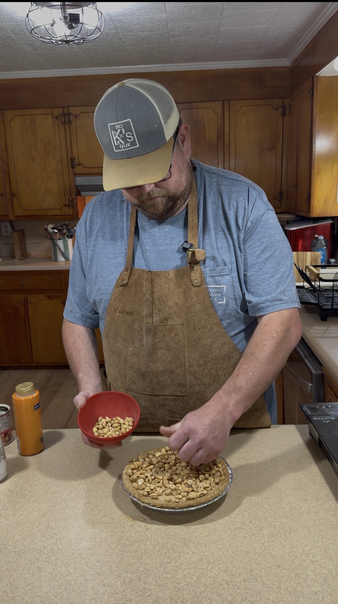 A man wearing a cap, glasses, and apron stands in a kitchen, sprinkling peanuts from a red bowl onto a pie crust on the counter. Wooden cabinets and kitchen utensils are visible in the background.