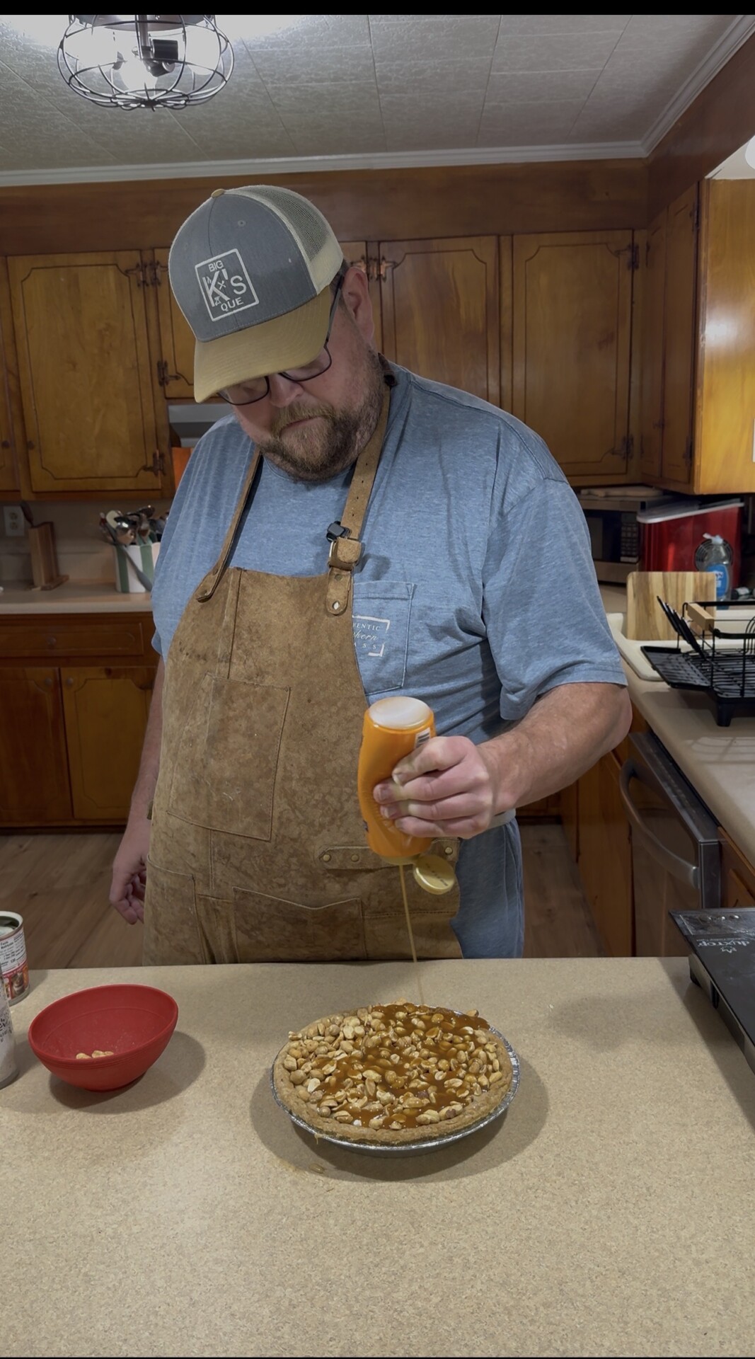 A man wearing an apron and cap pours a golden syrup from a bottle onto a pie topped with nuts in a kitchen with wooden cabinets. A red bowl and can sit on the counter nearby.