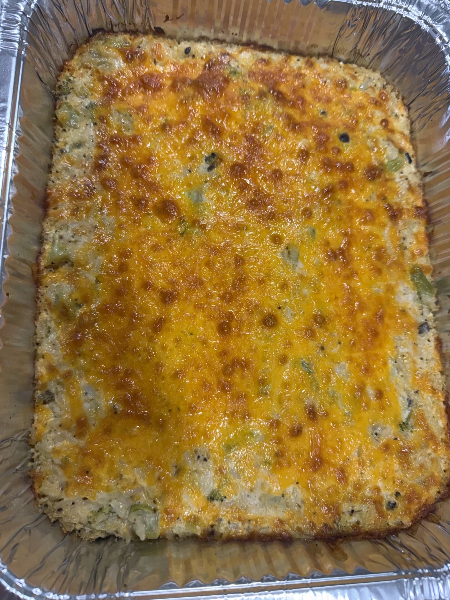 A freshly baked casserole in a rectangular aluminum tray, topped with a golden-brown melted cheese layer and specks of green vegetables are visible throughout the dish.