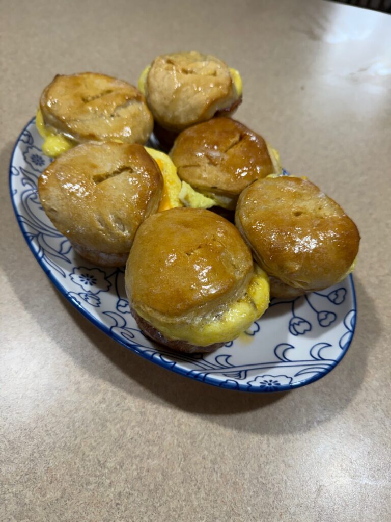 Six golden-brown biscuit sandwiches filled with egg are arranged on a white and blue floral-patterned plate, set on a beige countertop.