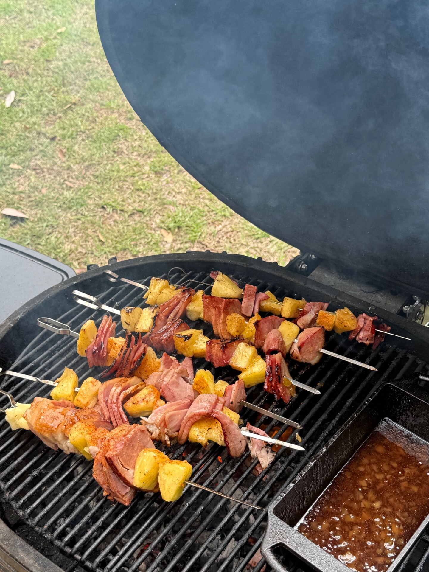 Skewers with chunks of ham and pineapple are being grilled on a barbecue with the lid open. Smoke rises, and a tray of sauce or marinade sits nearby. Grass is visible in the background.