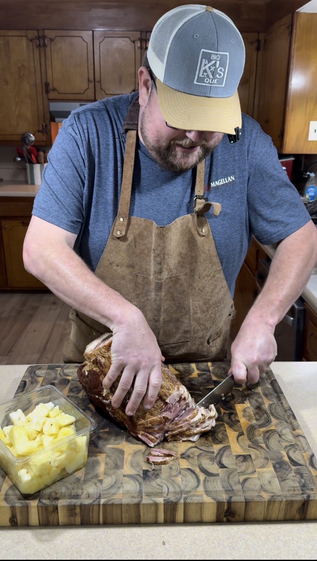A man wearing an apron and cap is slicing a large piece of cooked meat on a wooden cutting board in a kitchen. A glass container of pineapple chunks is next to the meat.