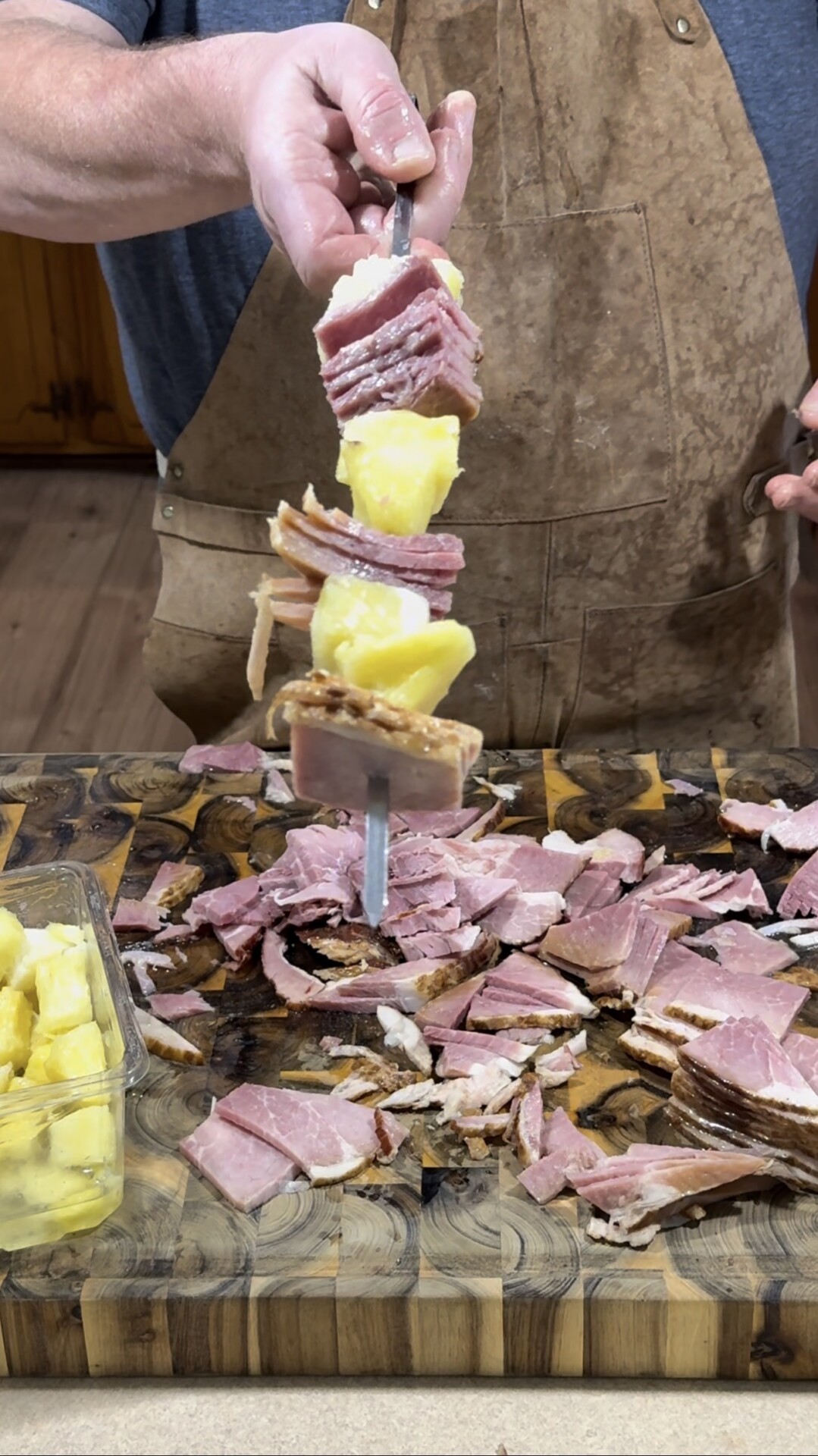 A person wearing a brown apron holds a skewer stacked with sliced ham, pineapple chunks, and bread over a cutting board covered with more ham slices and a container of pineapple chunks.