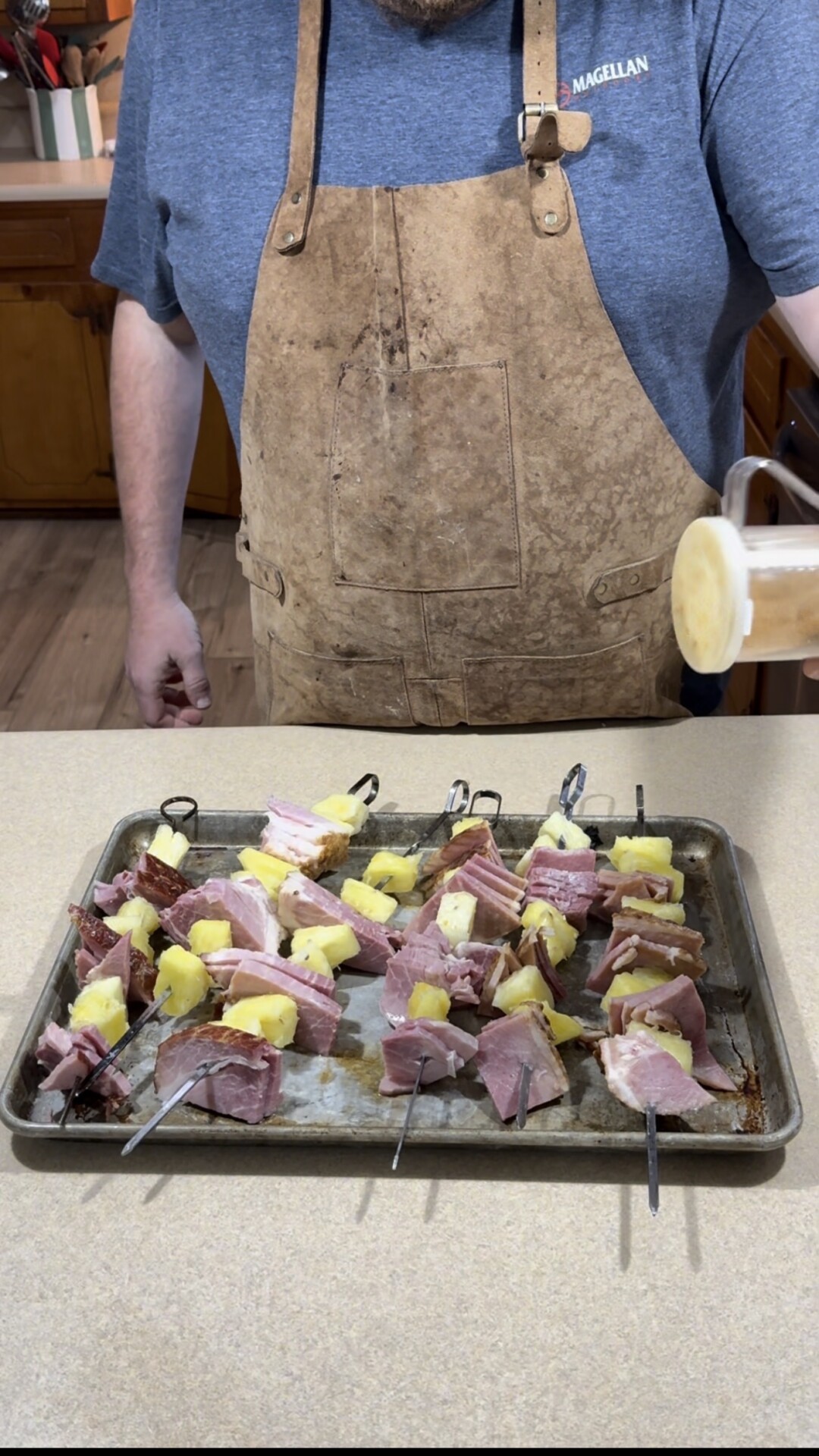 A person wearing a brown apron stands beside a baking sheet filled with skewers holding pieces of ham and pineapple, ready to cook.