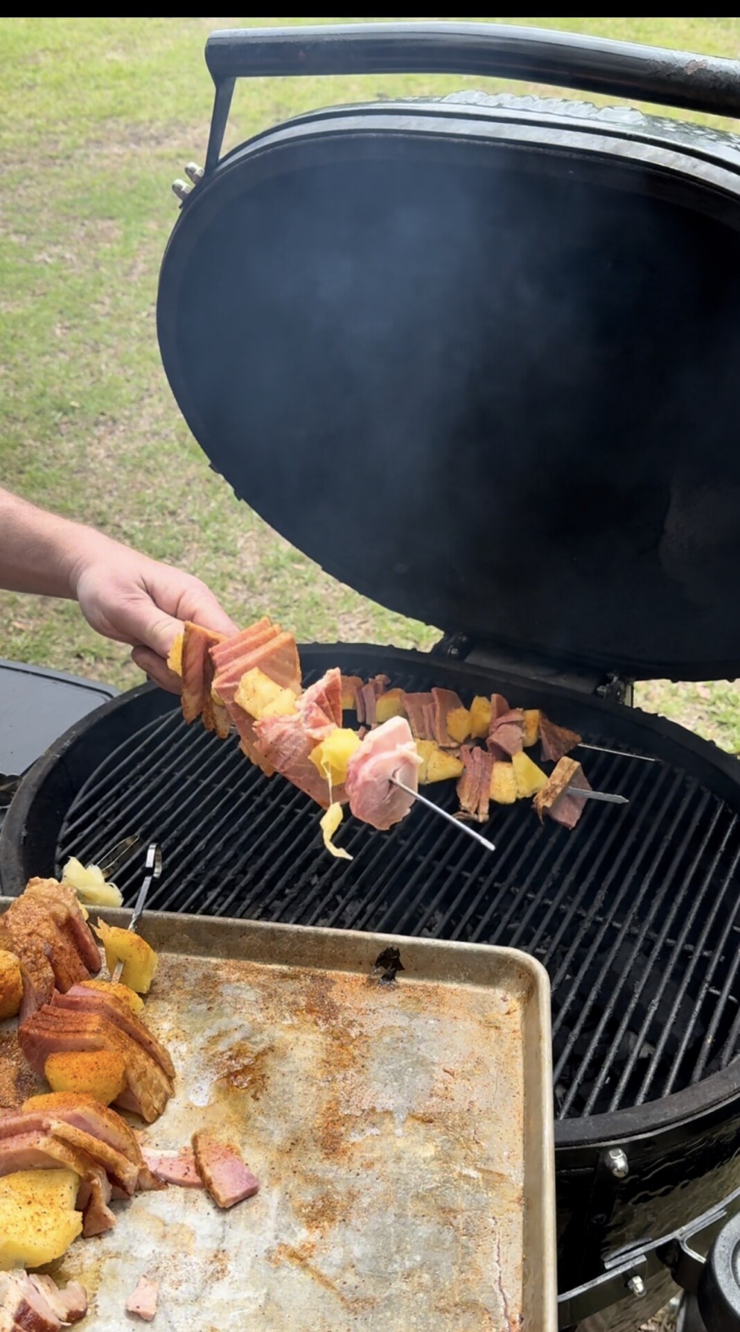 A person holds meat and pineapple skewers over an open grill, preparing to cook them. The grill lid is open, with smoke rising, and a tray with more uncooked skewers rests nearby. Grass is visible in the background.