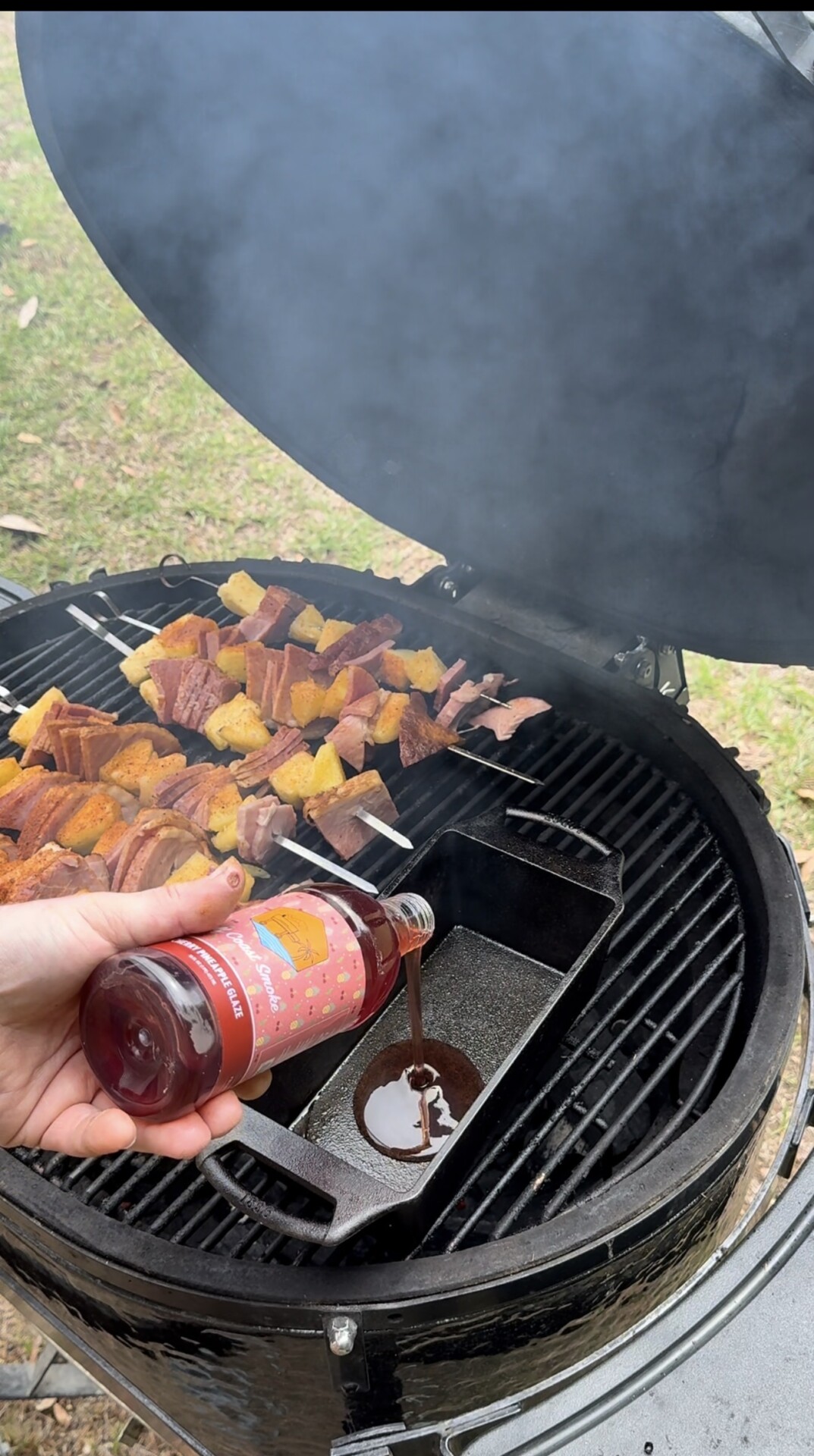 A hand pours BBQ sauce from a bottle into a pan on a grill filled with skewers of meat and vegetables. Smoke rises from the grill, and the scene is set outdoors.