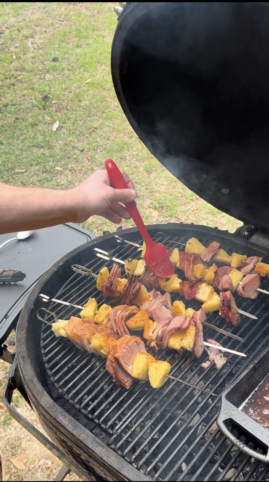 A person uses a red brush to glaze skewers of ham and pineapple on a round charcoal grill outdoors, with smoke rising and green grass in the background.
