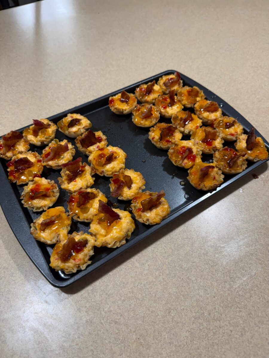 A black tray on a countertop holds about two dozen small appetizer bites topped with melted cheese and pieces of bacon, sprinkled with a red sauce or glaze.