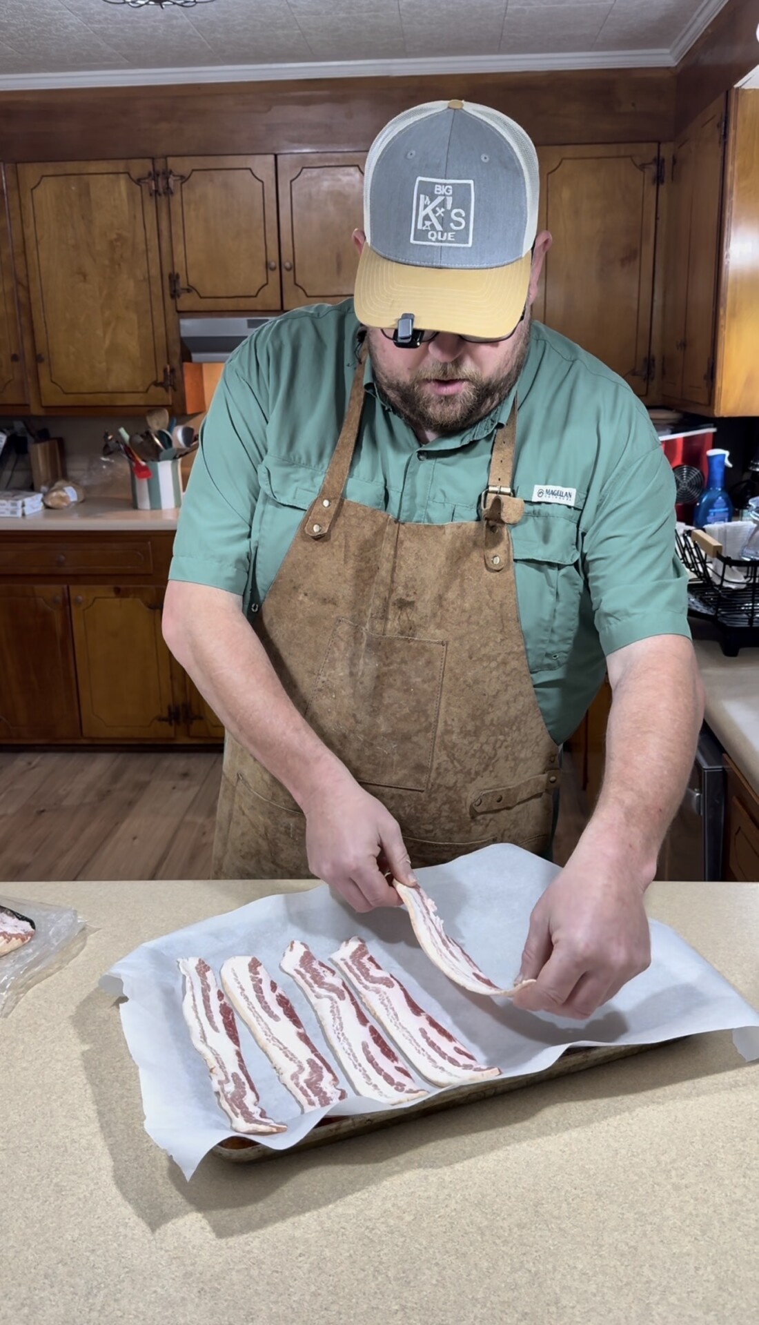 A man wearing an apron and a cap lays strips of raw bacon on parchment paper in a kitchen with wooden cabinets.