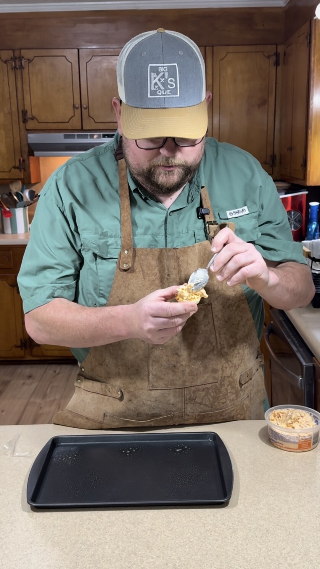 A man in a green shirt and brown apron stands in a kitchen, scooping a spread from a container onto a piece of bread over a baking tray on the counter.