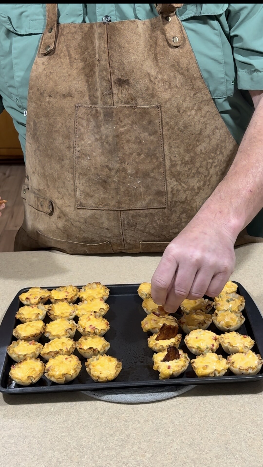 A person wearing a brown apron and green shirt is placing mini cheesy tarts or quiches onto a tray filled with similar baked appetizers on a kitchen counter.