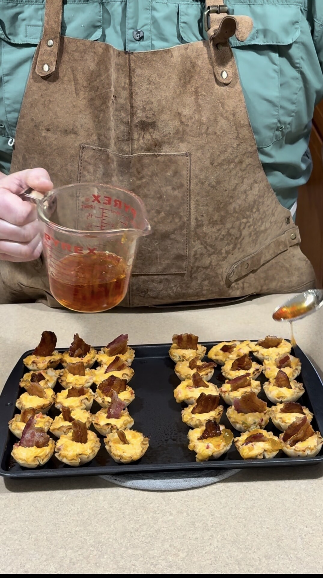 A person in a brown apron pours sauce from a measuring cup onto bite-sized savory tarts with bacon pieces, arranged in rows on a baking tray.