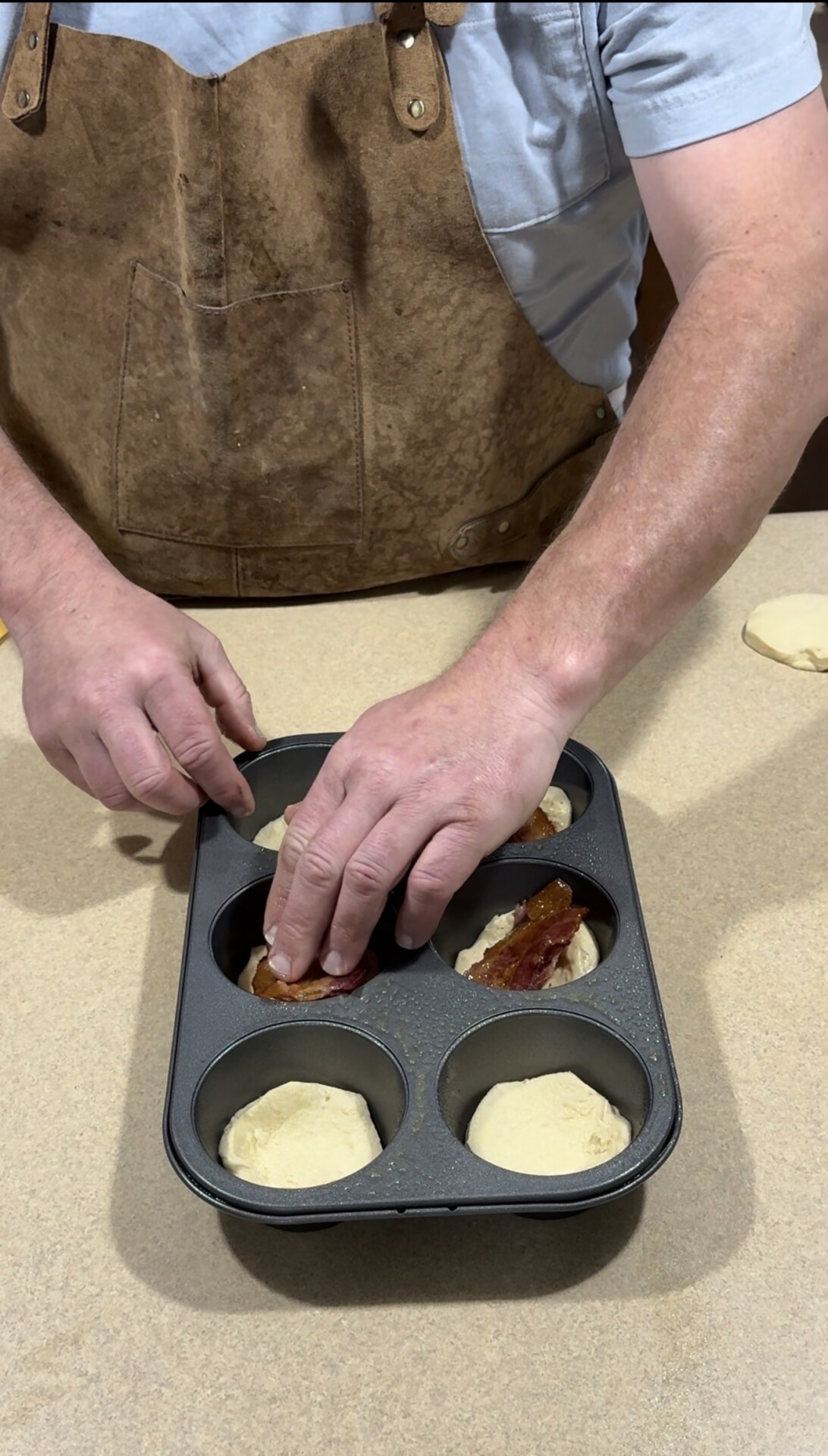 A person wearing a brown apron presses bacon into biscuit dough placed in a muffin tin on a beige countertop.
