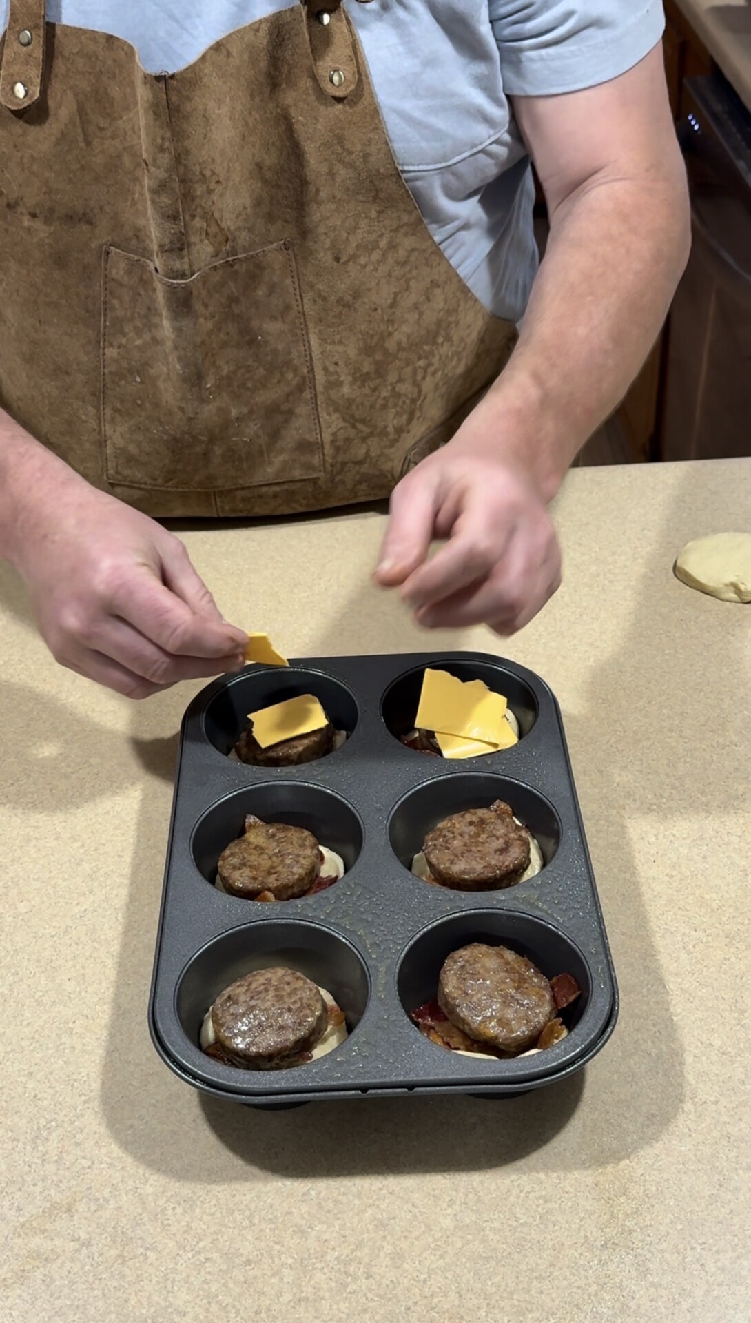 A person wearing a brown apron places pieces of sliced cheese into a muffin tin containing sausage patties and dough, preparing a savory dish on a kitchen counter.