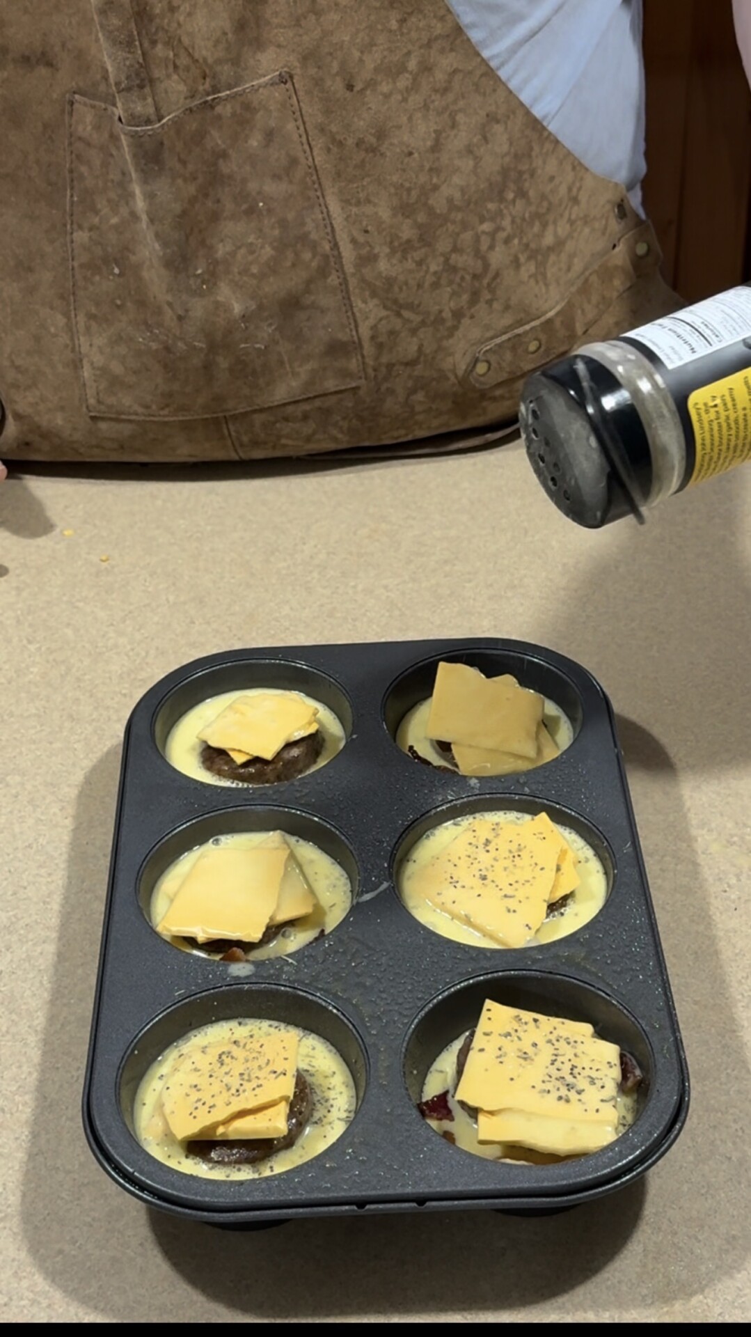 A person sprinkles seasoning on a muffin tin filled with egg mixture, meat, and cheese slices, preparing savory muffins on a kitchen counter.