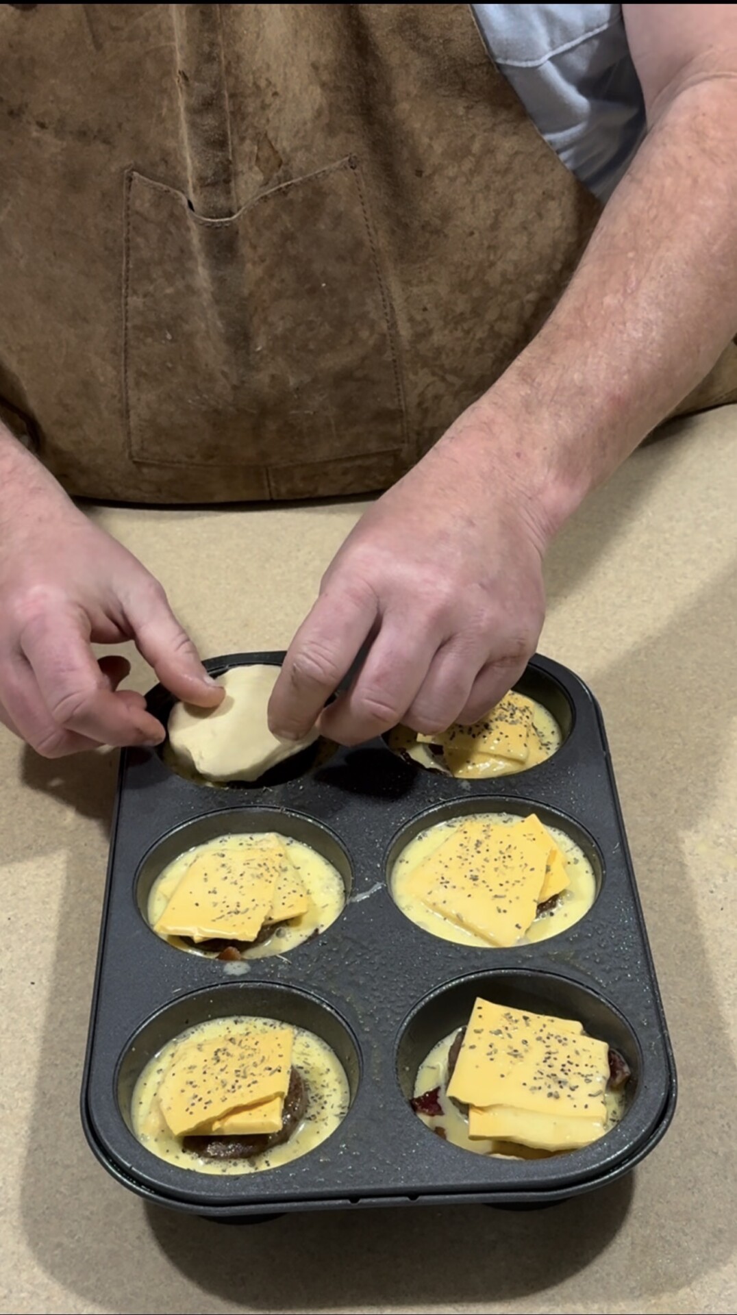 A person places dough over a muffin tin filled with egg mixture, cheese slices, and seasoning, preparing to bake individual servings. The person is wearing a brown apron.