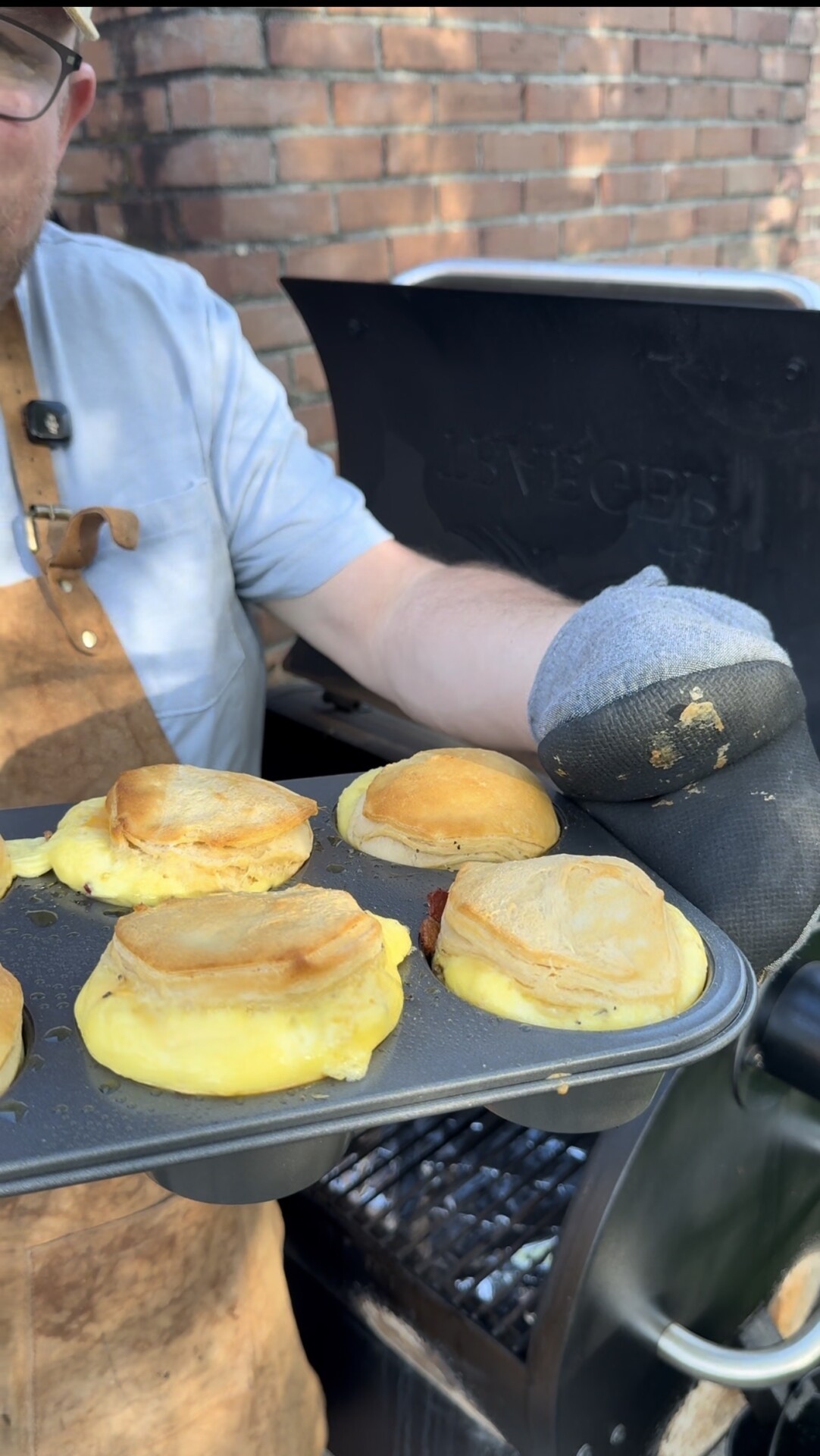 A person wearing an apron and oven mitt holds a muffin tin filled with breakfast sandwiches—biscuits with eggs and cheese—fresh off a grill in an outdoor setting.