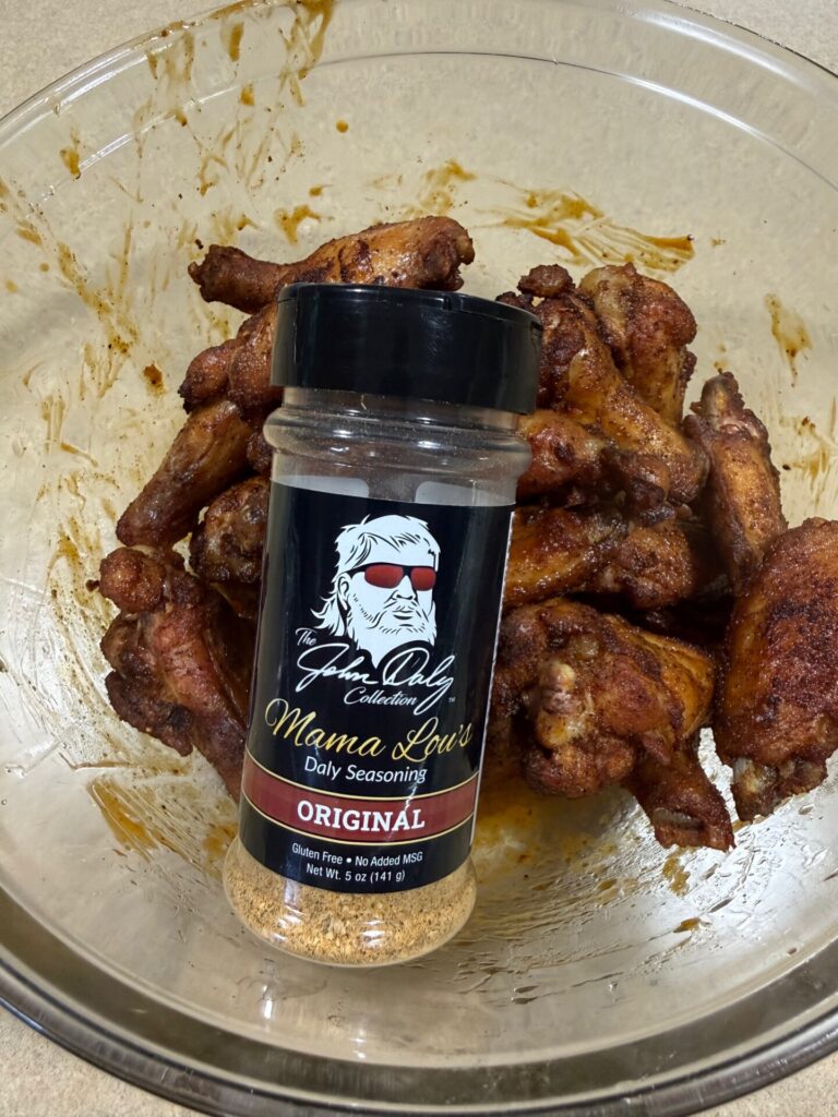 A bowl of cooked chicken wings with a jar of Mama Lou’s Daily Seasoning Original placed on top. The seasoning label features a drawing of a woman with sunglasses and white hair.