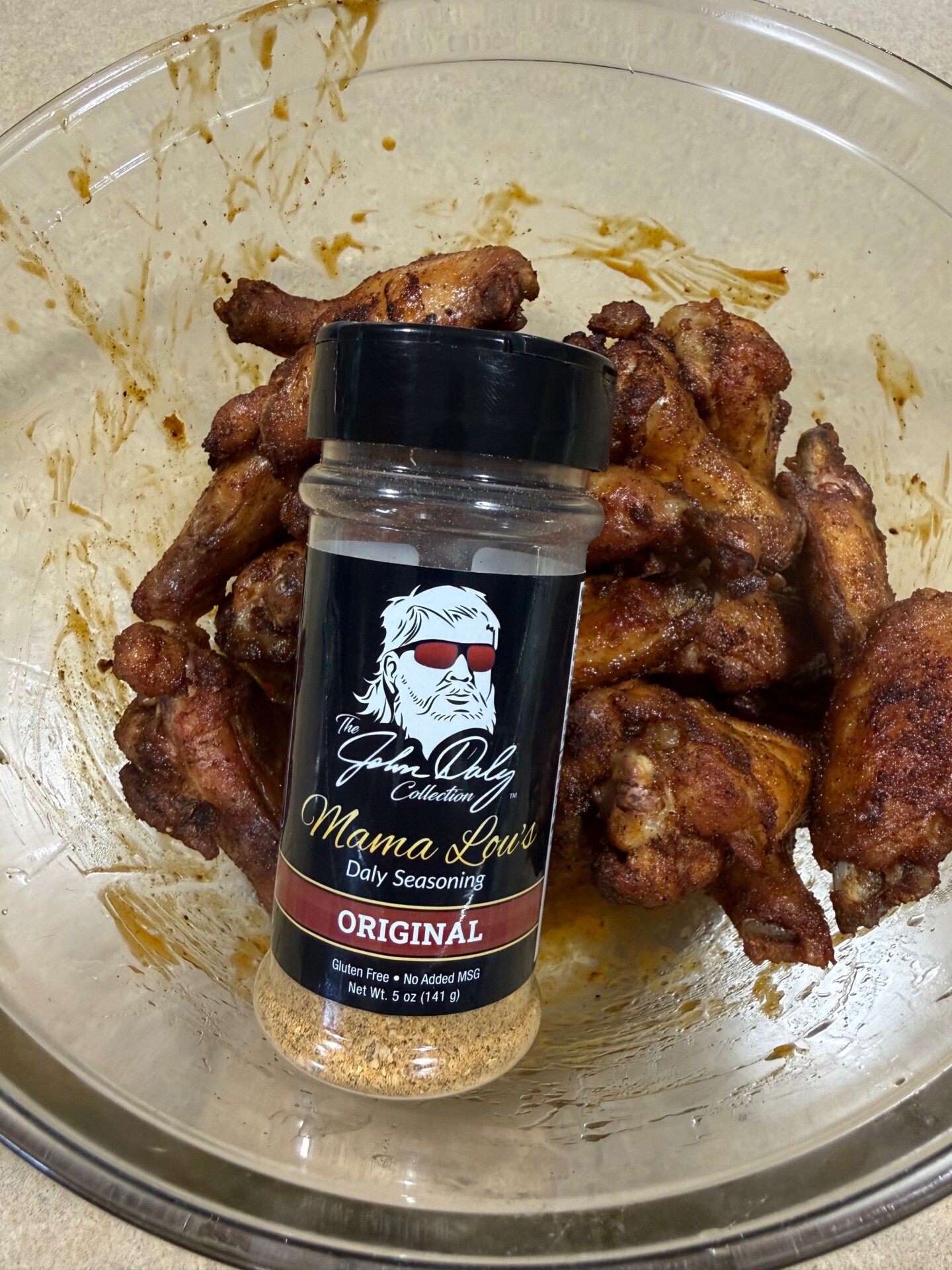 A bowl of cooked chicken wings with a jar of Mama Lou’s Daily Seasoning Original placed on top. The seasoning label features a drawing of a woman with sunglasses and white hair.